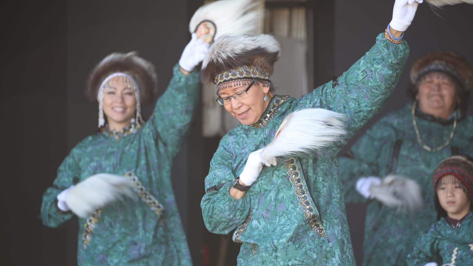 Alaska Native dancers in green regalia and fur headdresses performing with dance fans.